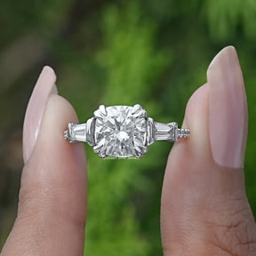 Front View Of A Cushion Cut Moissanite Three Stone Engagement Ring In Two Fingers