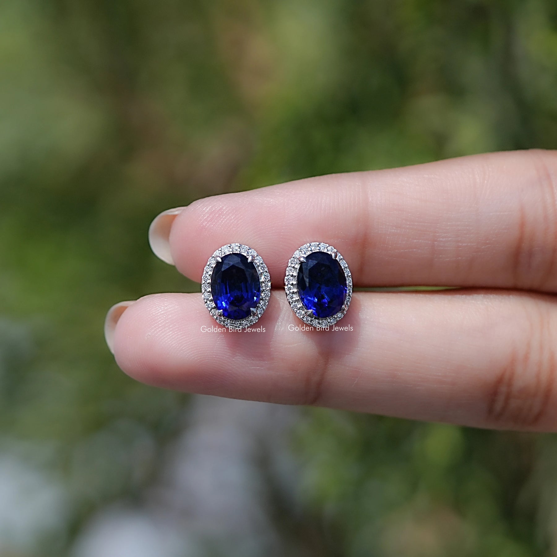 Blue gemstone earrings held between fingers with a blurred natural background